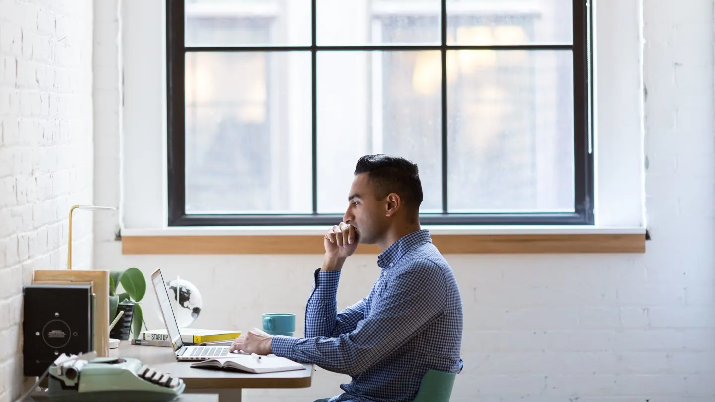A man at his desk by a window, hand to his chin, thinking as he looks at his laptop
